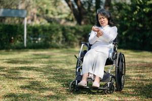 asiático antiguo mujer sentado en un silla de ruedas al aire libre en el parque tener dolor en el brazos, muñecas y cuerpo en Dom ligero foto