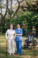 Asian careful caregiver or nurse and the happy patient in a wheelchair are walking in the garden to help and encourage and rest your mind with green nature. Help support yourself to learn to walk. photo