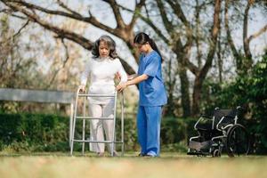 Asian careful caregiver or nurse and the happy patient in a wheelchair are walking in the garden to help and encourage and rest your mind with green nature. Help support yourself to learn to walk. photo