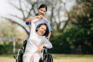 Asian careful caregiver or nurse and the happy patient in a wheelchair are walking in the garden to help and encourage and rest your mind with green nature. Help support yourself to learn to walk. photo