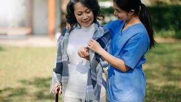 Asian careful caregiver or nurse and the happy patient in a wheelchair are walking in the garden to help and encourage and rest your mind with green nature. Help support yourself to learn to walk. photo