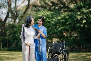 Asian careful caregiver or nurse and the happy patient in a wheelchair are walking in the garden to help and encourage and rest your mind with green nature. Help support yourself to learn to walk. photo