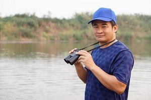 Asian man wears blue cap, hold binocular at the lake, nature source. Concept, nature exploration. Ecology study.  Pastime activity, lifestyle. Man explore environment photo