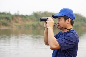 Asian man wears blue cap, hold binocular at the lake, nature source. Concept, nature exploration. Ecology study.  Pastime activity, lifestyle. Man explore environment photo