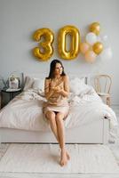 contento mujer sostiene un cumpleaños pastel con un vela en su manos, sentado en el cama en el dormitorio decorado para el fiesta y teniendo divertido foto