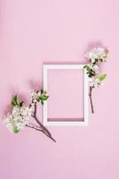 White blank frame with copy space lies on a pink background surrounded by blossoming branches of an apple tree. Spring postcard concept. Top view, flat lay photo