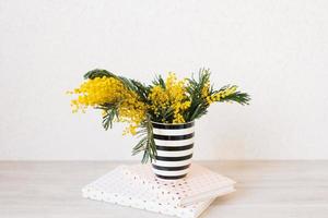 Branches of mimosa flowers stand in a black and white striped vase on notebooks on the table photo