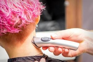 Back view of hairdresser's hand shaving nape and neck with electric trimmer of young caucasian woman with short pink hair in beauty salon. photo