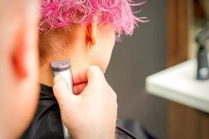 Back view of hairdresser's hand shaving nape and neck with electric trimmer of young caucasian woman with short pink hair in beauty salon. photo