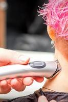 Back view of hairdresser's hand shaving nape and neck with electric trimmer of young caucasian woman with short pink hair in beauty salon. photo