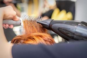 Closeup of master's hand with blow-drying and hairbrush blowing female red hair in a salon. photo