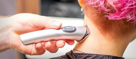 Back view of hairdresser's hand shaving nape and neck with electric trimmer of young caucasian woman with short pink hair in beauty salon. photo