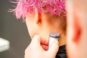 Back view of hairdresser's hand shaving nape and neck with electric trimmer of young caucasian woman with short pink hair in beauty salon. photo