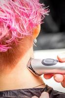 Back view of hairdresser's hand shaving nape and neck with electric trimmer of young caucasian woman with short pink hair in beauty salon. photo