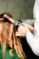 Hairdresser using curling tongs curls long brown hair on the young caucasian girl in a beauty salon. photo