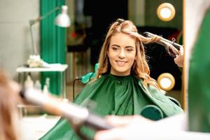 Hairdresser using curling tongs curls long brown hair on the young caucasian girl in a beauty salon. photo