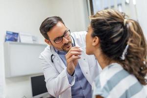 A young woman sits on an exam table across from her doctor. The doctor reaches forward with a tongue depressor as the woman looks up and sticks out her tongue. photo