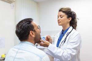 Young man sits on an exam table across from his doctor. The doctor reaches forward with a tongue depressor as the man looks up and sticks out his tongue. photo