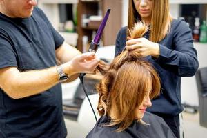 Two hairstylists using curling iron on customers long brown hair in a beauty salon. photo