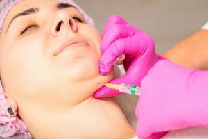 The cosmetologist makes lipolytic injection on the chin of a young woman against the double chin in a beauty salon. photo