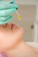 Close up of beautician holding pipette and applying oil serum drop on the face of a young woman in spa salon. photo