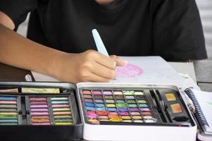 Asian boy is spending his free times during his summer vacation in the living room by drawing, painting and sketching with watercolor dry bar in tray in front of him, soft focus. photo