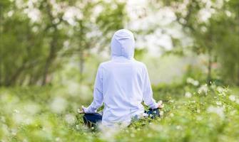 Rear view of woman in hoodie is relaxingly practicing meditation yoga in forest full of daisy flower in summer to attain happiness from inner peace wisdom with morning light for healthy mind and soul photo