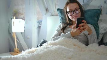 Woman in glasses lies in a bed and using a smartphone before bedtime video