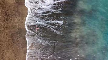 Aerial view of a man in red shorts rotates around himself stretching arms to the sides on the beach with black sand video