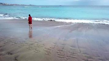 Aerial view of a man in red shorts rotates around himself stretching arms to the sides on the beach with black sand video