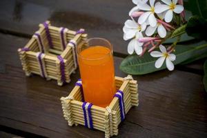 A glass of orange juice with water glass holder made of bamboo sticks and fiber. Decorative wooden table with Beautiful Plumeria flowers. Bamboo Glass Holder. photo