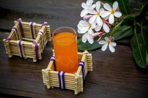 A glass of orange juice with water glass holder made of bamboo sticks and fiber. Decorative wooden table with Beautiful Plumeria flowers. Bamboo Glass Holder. photo