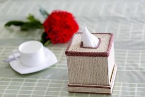 Raw fiber color handmade Napkin box on the dining table with a red flower and a white tea-cup. Tissue paper box on restaurant. photo