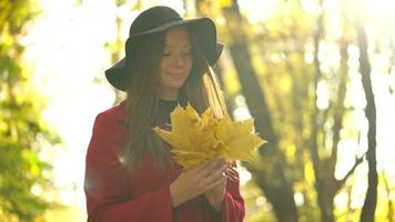 Beautiful girl sits in the autumn forest and collects a bouquet of yellow maple leaves. Slow motion video