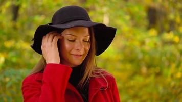 Portrait of a beautiful smiling girl in a black hat with a yellow maple leaf in the foreground in the autumn forest. Slow motion video