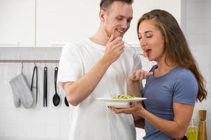 joven Pareja saboreo un ensalada mientras en pie en cocina. contento hombre y mujer comiendo ensalada juntos a hogar foto