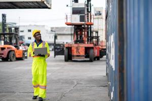 African technician dock worker in protective safety jumpsuit uniform and with hardhat and use digital tablet at cargo container shipping warehouse. transportation import,export logistic industrial photo