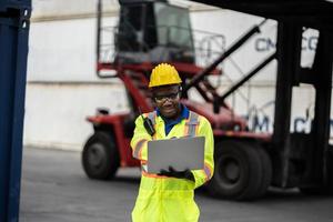 African technician dock worker in protective safety jumpsuit uniform and with hardhat and use digital tablet at cargo container shipping warehouse. transportation import,export logistic industrial photo