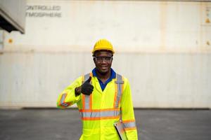 retrato de africano contento trabajador en protector la seguridad mono uniforme con casco de seguridad y utilizar ordenador portátil computadora a carga envase Envío depósito. transporte importación y exportación logístico industrial foto