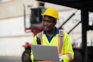 African technician dock worker in protective safety jumpsuit uniform and with hardhat and use digital tablet at cargo container shipping warehouse. transportation import,export logistic industrial photo