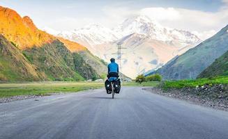 masculino persona en azul chaqueta ciclismo con turismo bicicleta rodeado por montañas y verde verano naturaleza. excursión alrededor Kazbegi nacional parque. Georgia viaje ciclismo Días festivos concepto foto