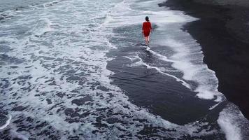 aéreo ver de un niña en un rojo vestir caminando en el playa con negro arena. tenerife, canario islas, España video