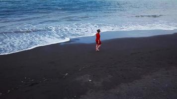 aereo Visualizza di un' ragazza nel un' rosso vestito a piedi su il spiaggia con nero sabbia. tenerife, canarino isole, Spagna video