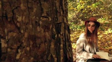 hermosa mujer en un sombrero y Gafas de sol es acostado en un tartán en un otoño parque o bosque, descansando desde el bullicio de el ciudad y disfruta bueno clima. lento movimiento video