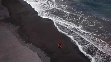 Antenne Aussicht von ein Mädchen im ein rot Kleid Gehen auf das Strand mit schwarz Sand. Teneriffa, Kanarienvogel Inseln, Spanien video