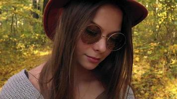 Portrait of a beautiful smiling girl in a hat and sunglasses with a yellow maple leaf in the foreground in the autumn forest. Slow motion video