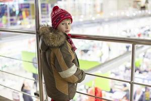 A boy of elementary age in winter clothes is standing by a metal handrail indoors. photo