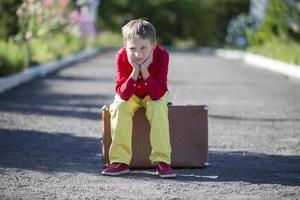 A sad boy sits on an old suitcase on the road. The child is about to leave. photo