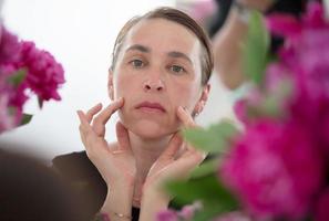 un medio Envejecido mujer examina arrugas en contra un antecedentes de flores cuidado para envejecimiento piel. foto