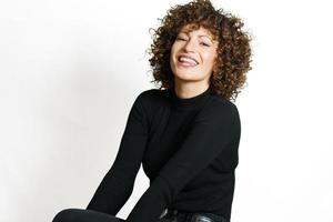 Smiling woman with curly hair sitting against white wall photo